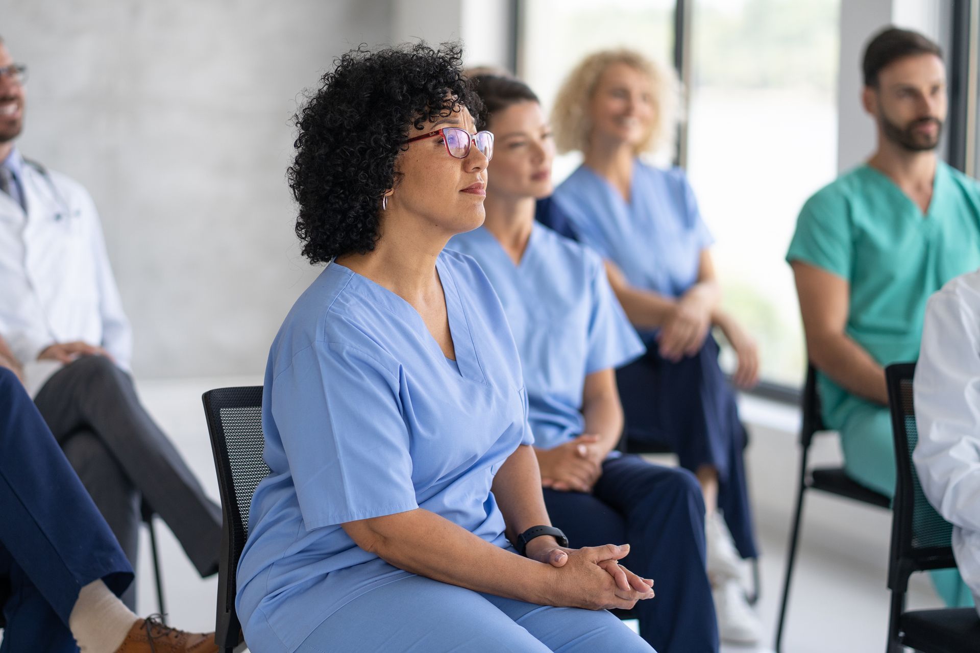 Medical professionals in scrubs and white coats sit attentively in chairs indoors.