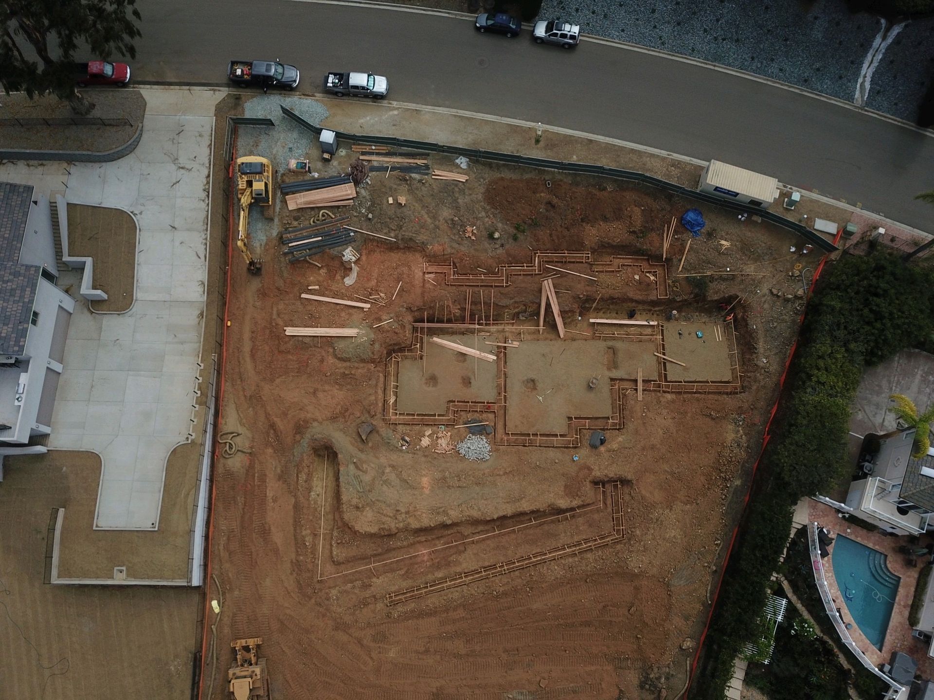 Aerial view of a construction site beside a curved road, with dirt foundation work and parked vehicles nearby.
