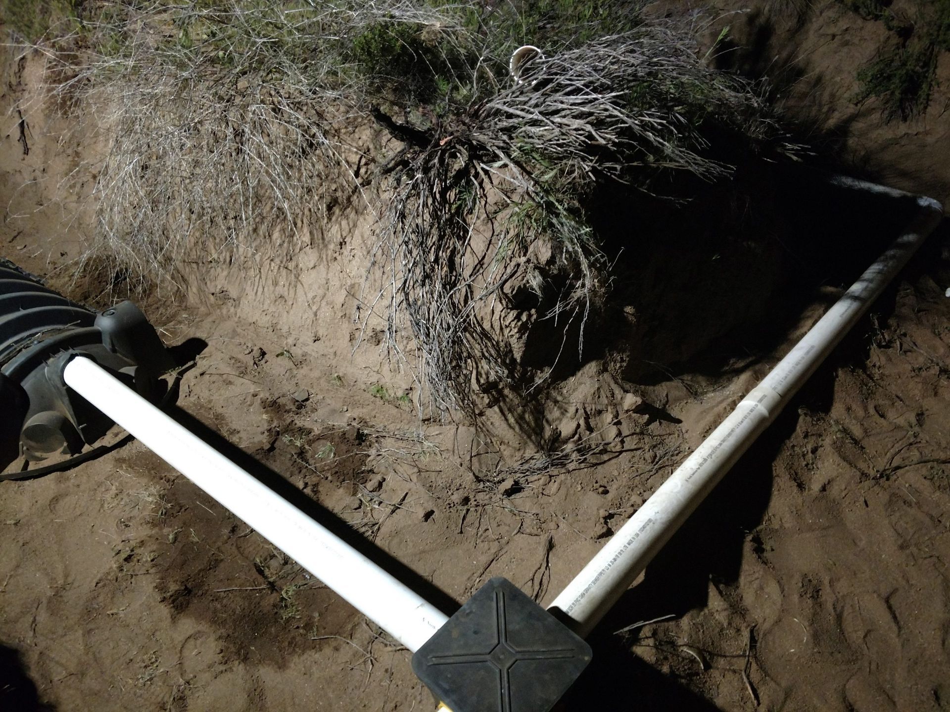 White PVC pipes forming a Y-shape in a muddy trench beside an exposed dirt bank.