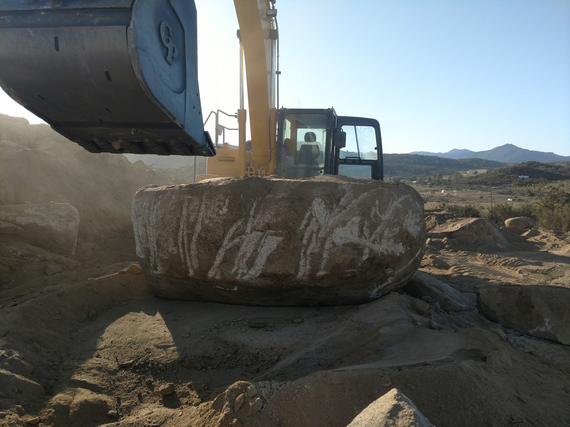 Excavator lifting a large boulder at a dusty construction site under a clear sky.