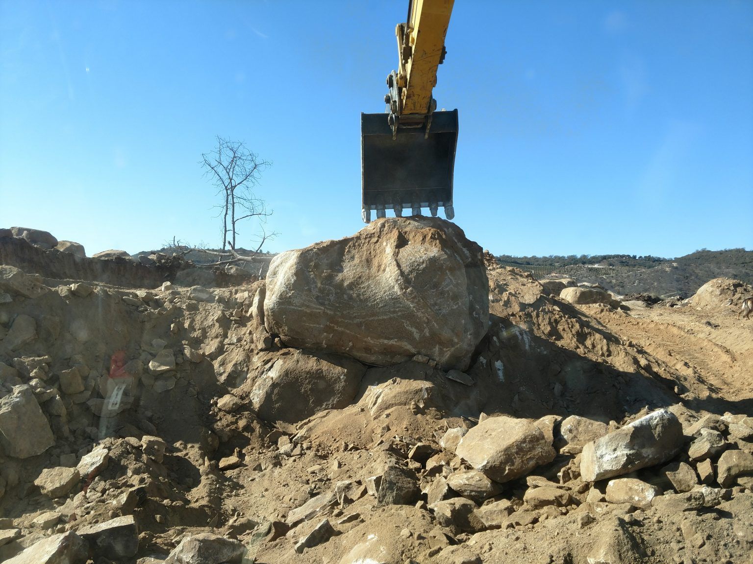 Excavator bucket lifting a large boulder at a rocky construction site under a clear blue sky
