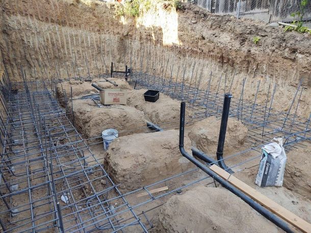 Foundation excavation with steel rebar grid, pipes, and concrete footings in a trench.