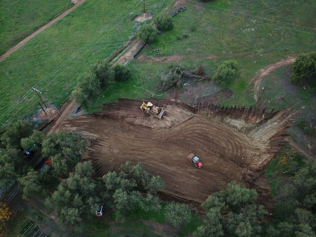 Aerial view of a muddy field with excavators and workers beside green farmland and trees