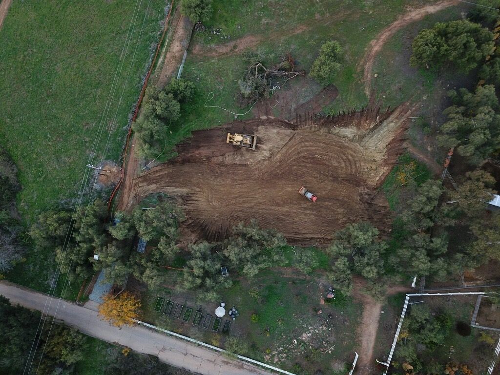 Aerial view of a dirt construction site with heavy machinery beside a grassy area and road