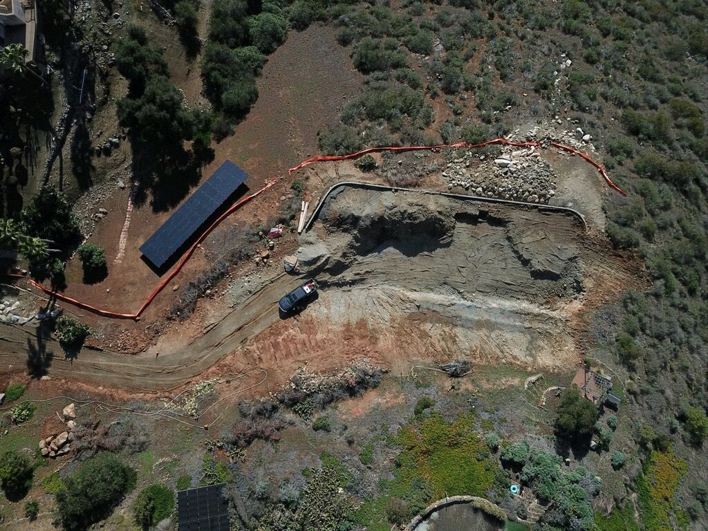 Aerial view of a construction site with an excavator, dirt road, and a small solar-roofed structure.