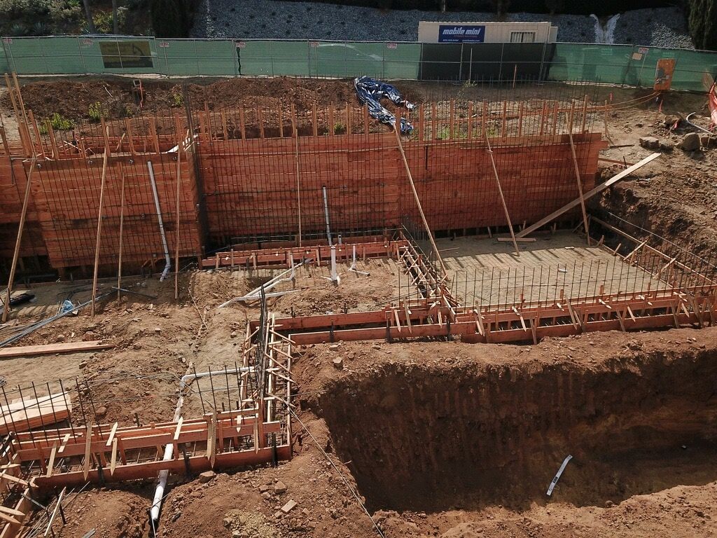 Construction site with large excavated pit, wooden formwork, and rebar on a dirt lot
