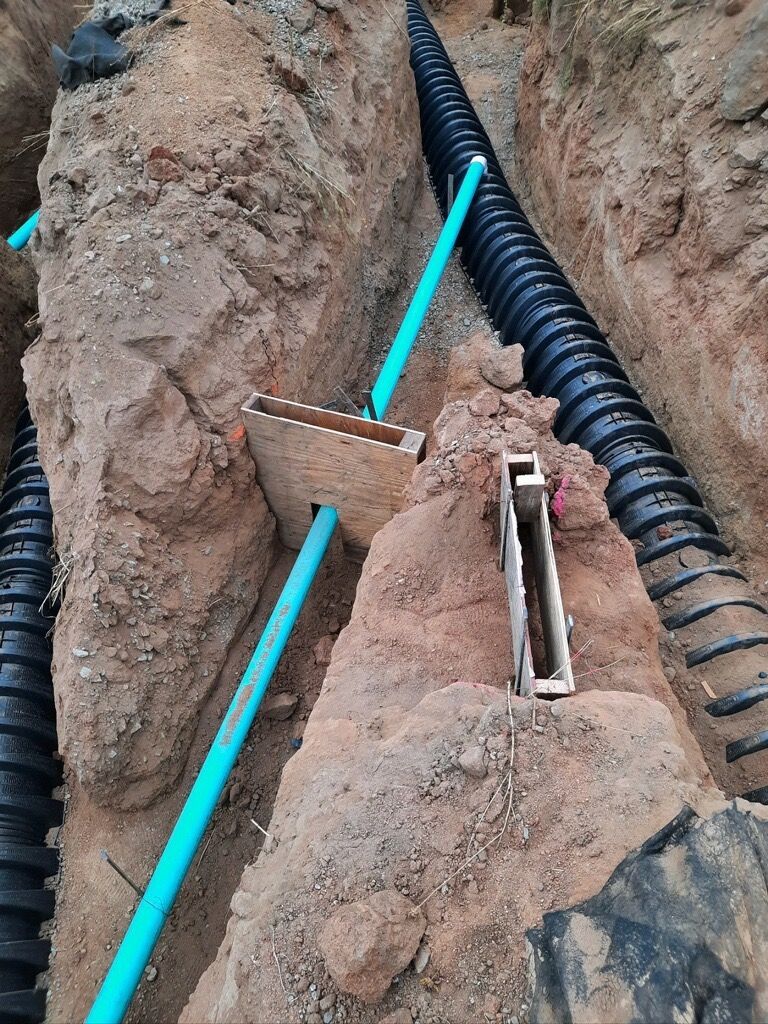 Construction trench with blue conduit pipes, wooden spacers, and a black corrugated pipe in dirt.
