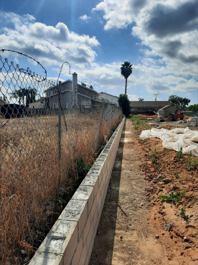 Concrete drainage channel beside a barbed-wire fence, with dry weeds, white-covered crops, and a cloudy sky