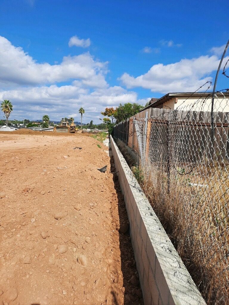Dirt road beside a chain-link fence under a bright blue sky with scattered clouds