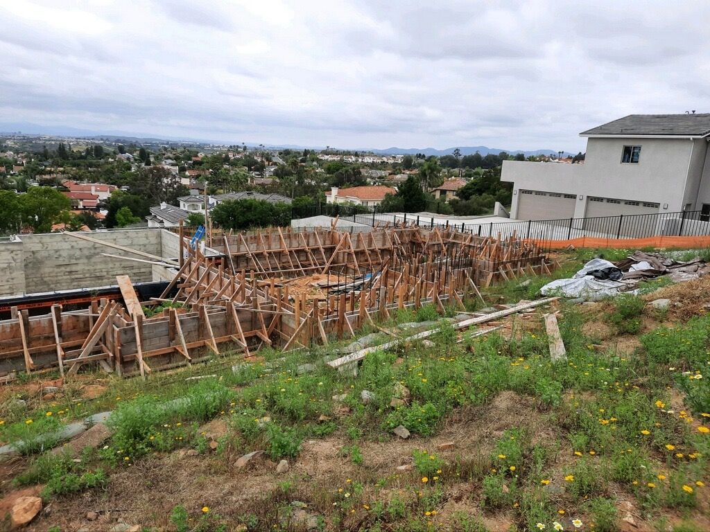 Construction site on a hillside with wooden formwork, overlooking a residential neighborhood under cloudy skies