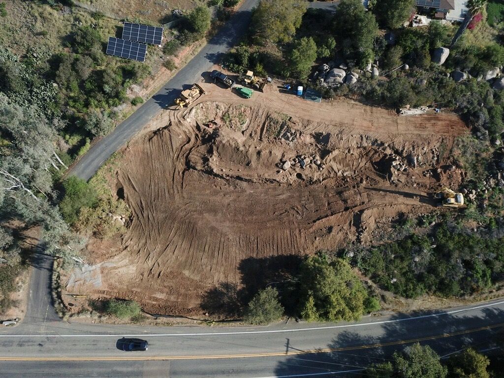 Aerial view of a large dirt landslide beside a road, with trees and a few parked vehicles nearby.