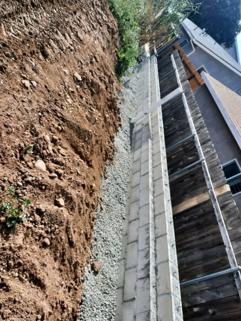 Steep dirt and gravel trench beside a house foundation with retaining wall and fence