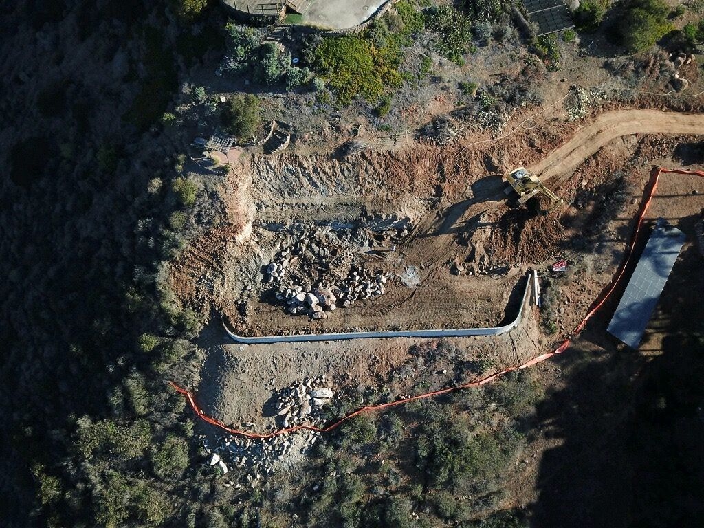 Aerial view of an excavated construction site with exposed soil, rocks, and a trench along the edge of trees.