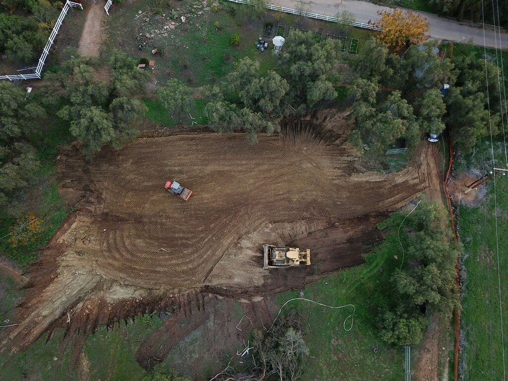 Aerial view of a dirt clearing surrounded by trees, with a small excavator and one person working.