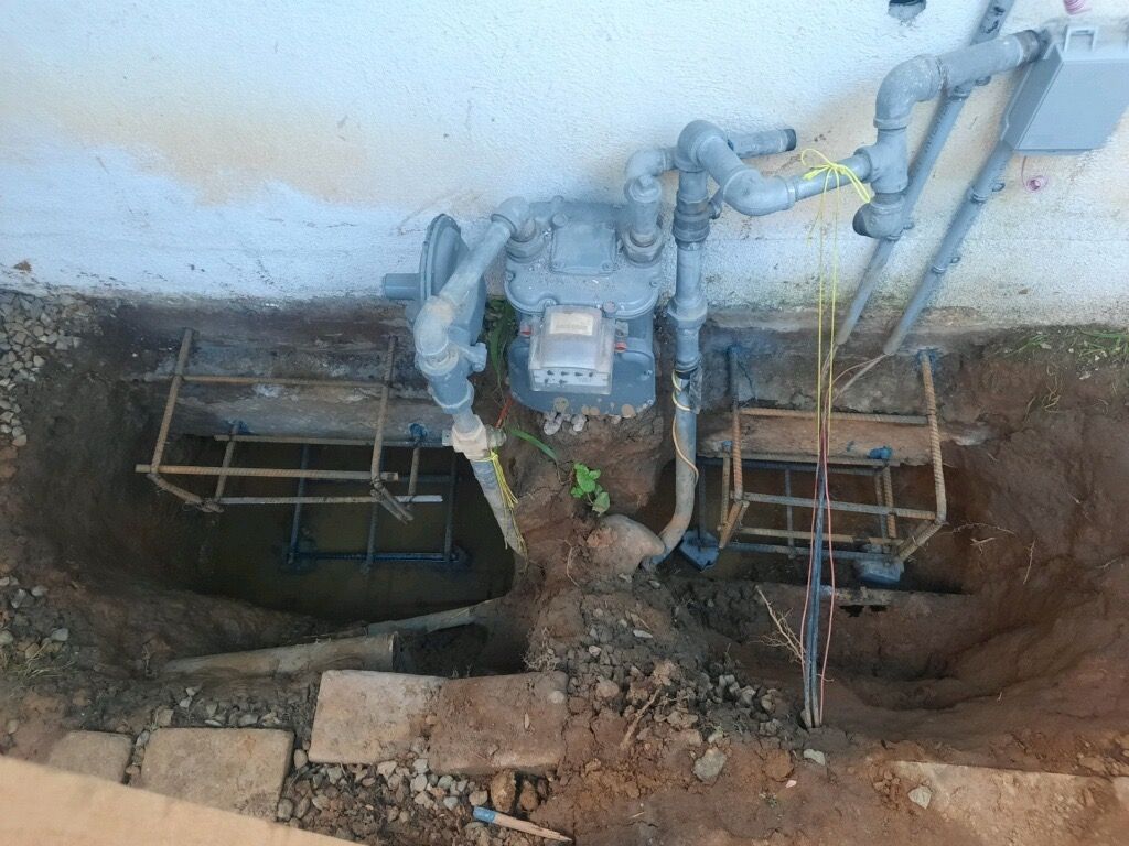 Open utility trench with gas meters and pipes against a wall, with exposed wiring and dirt around it