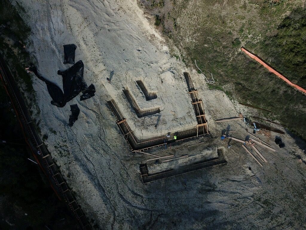 Aerial view of a construction site with concrete forms, dark equipment, and a dirt access path.