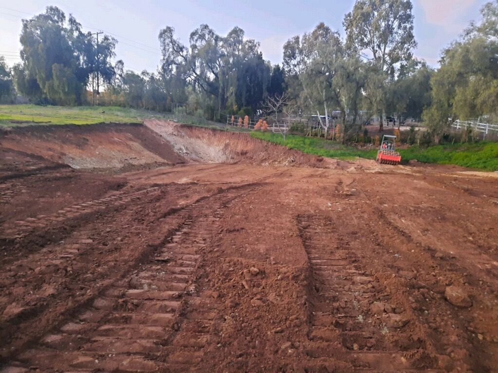 Dirt construction site with graded red soil, tire tracks, and trees under a cloudy sky