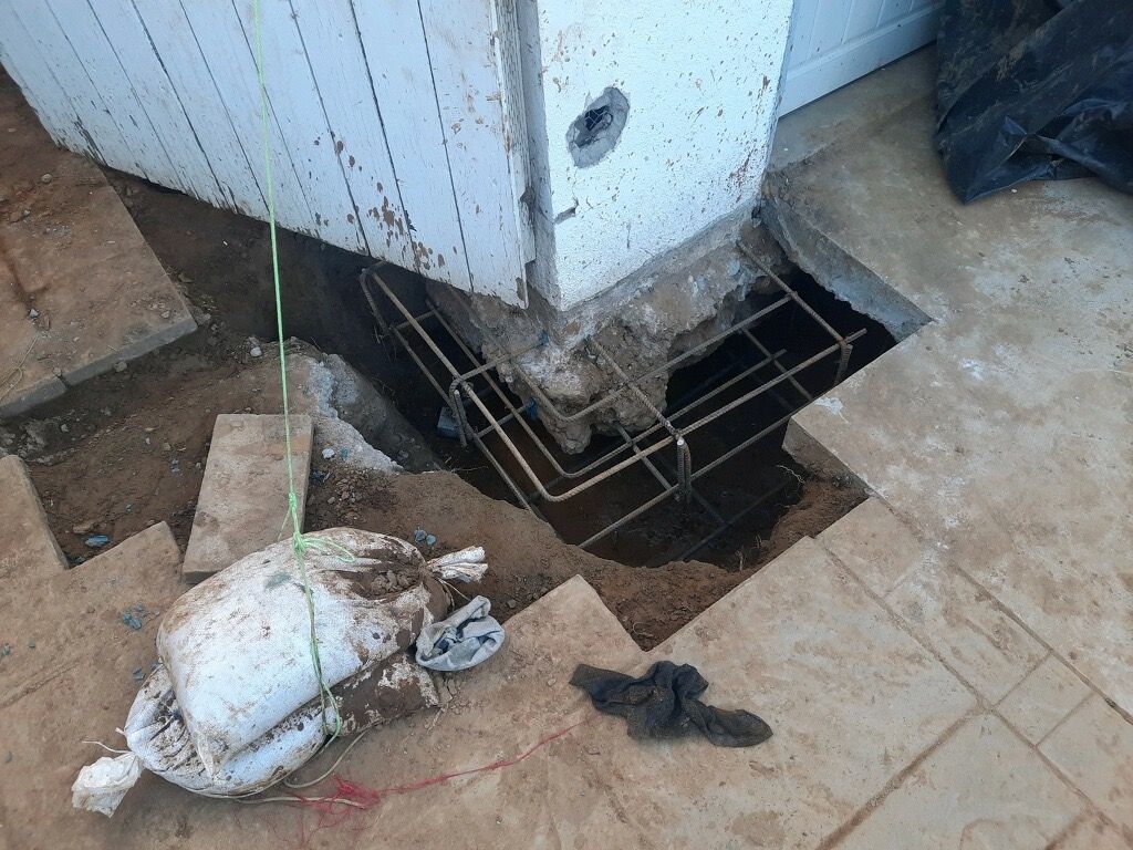 Damaged floor around a wall opening with exposed wiring and a fallen white bag in a dusty room
