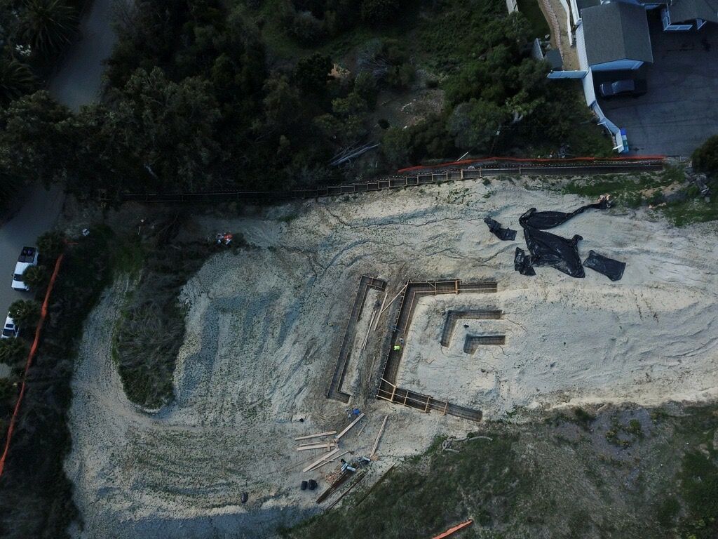 Aerial view of a sandy construction site with a large stepped concrete foundation and surrounding trees.