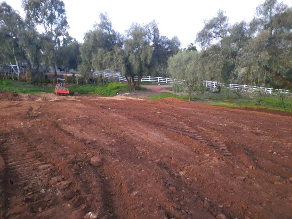 Dirt field with fresh tire tracks and a red tractor near trees under a cloudy sky