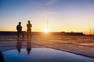 Two workers in safety vests on a rooftop, silhouetted against a sunrise.