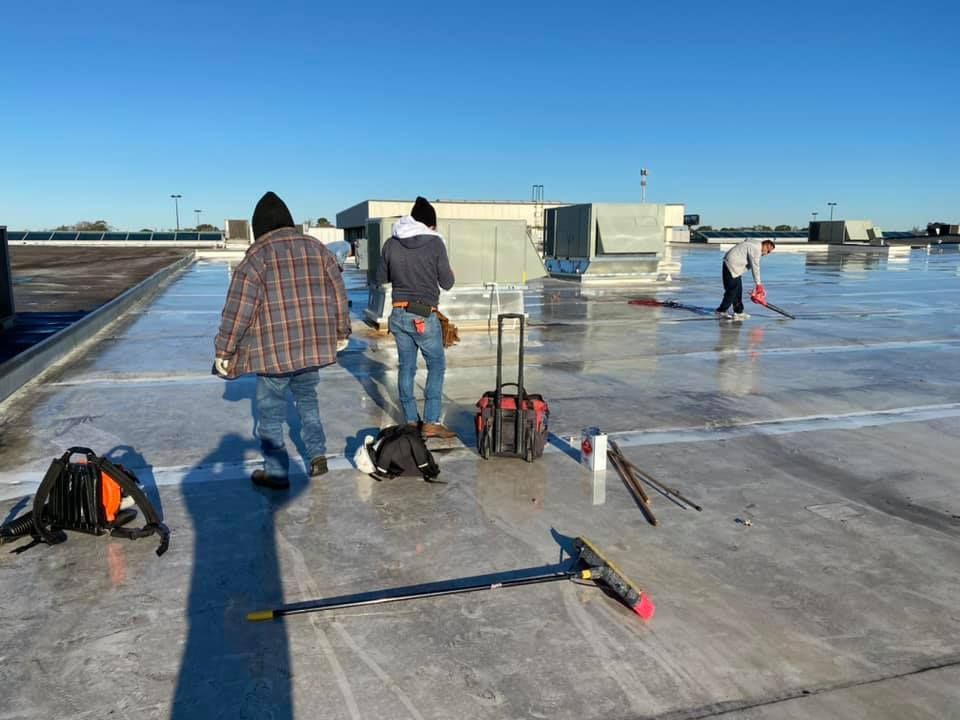 Workers on a flat, icy roof, applying sealant. Blue sky, tools, and a sunny day are in the image.