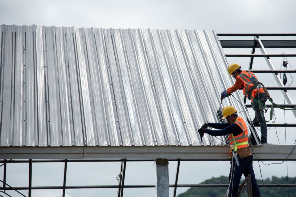 Two roofers in safety vests installing corrugated metal roofing on a metal frame structure.