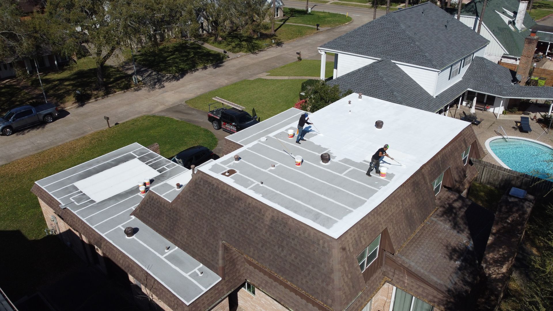 Two workers on a white roof applying sealant. Brown roofed house with a pool and street.