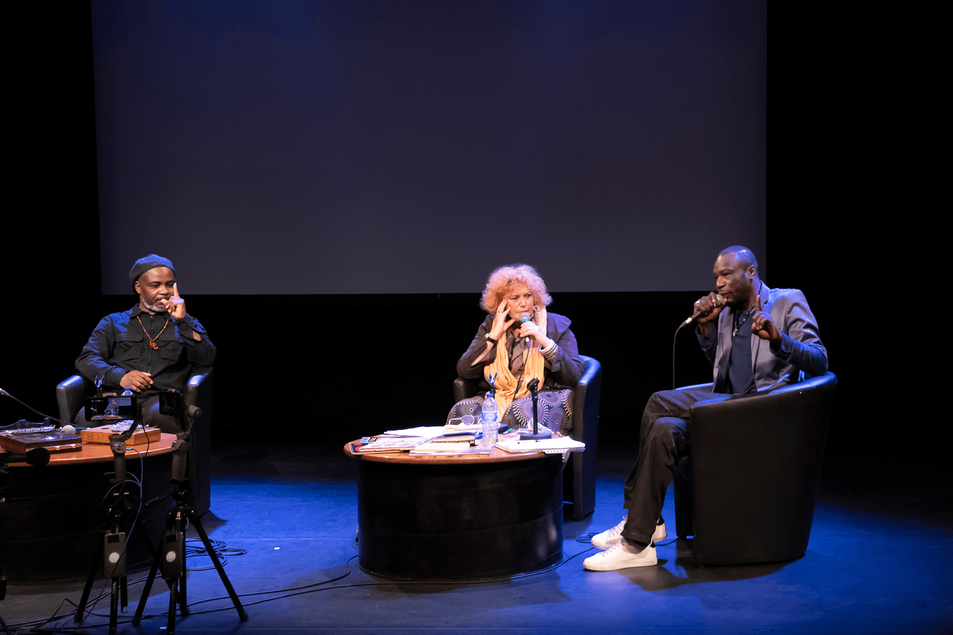 Trois personnes sont assises à une table sur une scène et parlent dans des microphones.