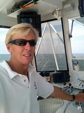 Captain Pete wearing sunglasses and a white polo shirt steers a boat from the bridge, looking toward the camera.