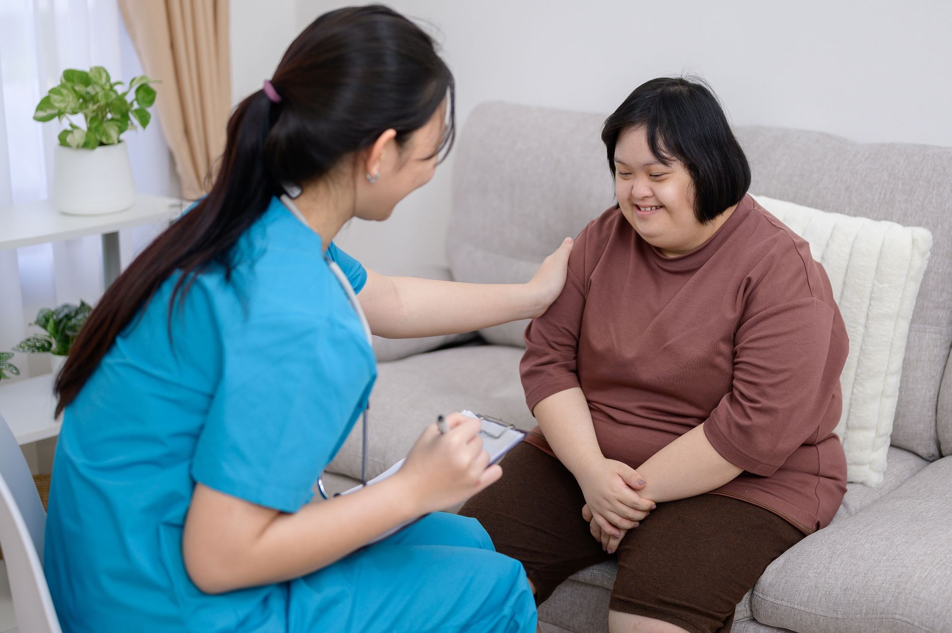 Nurse in blue scrubs speaking gently with a smiling patient on a sofa in a living room