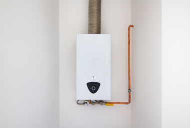 A white wall-mounted gas water heater with a metallic flue pipe and copper plumbing connections on a white wall.