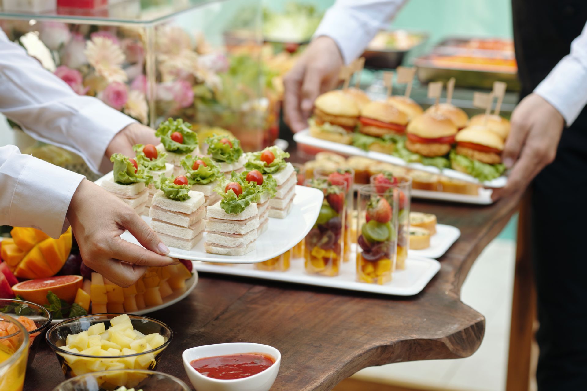 Catered buffet table with sandwiches, hors d’oeuvres, fruit, and desserts being served by gloved hands