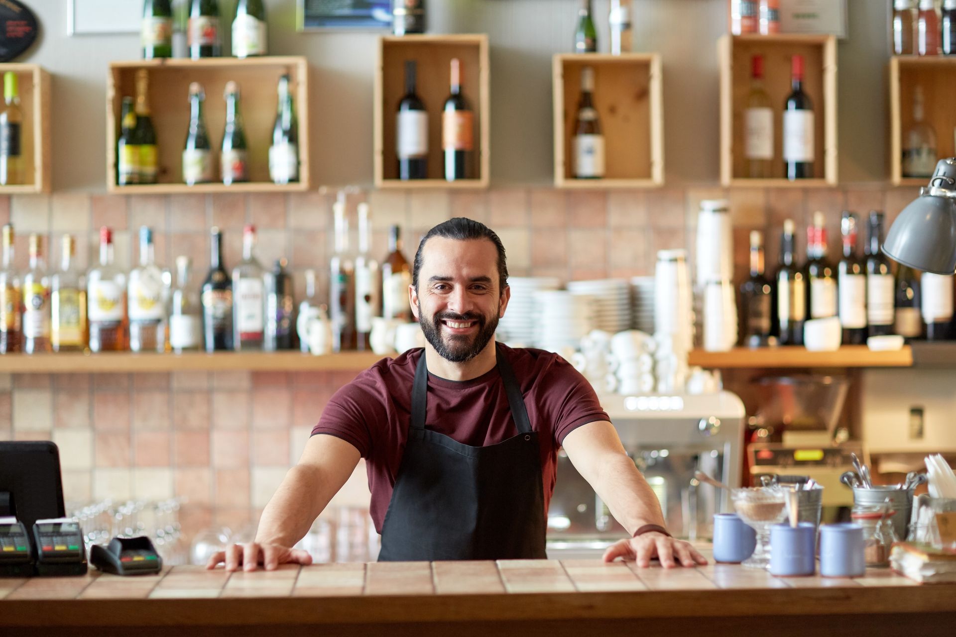 Smiling bartender behind a bar with shelves of bottles and glasses, wearing an apron.