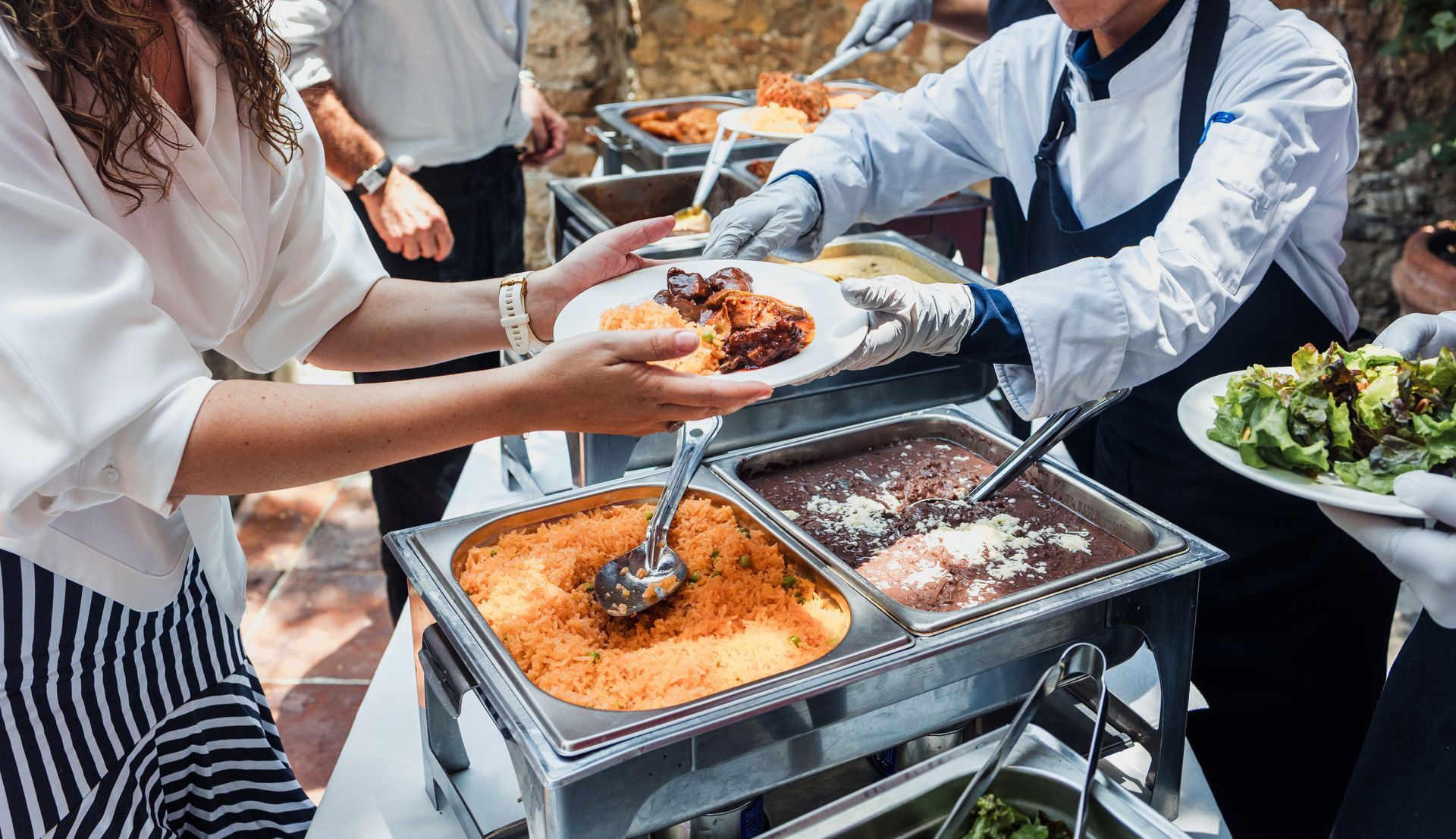 People serving buffet food with tongs from chafing dishes at an outdoor event