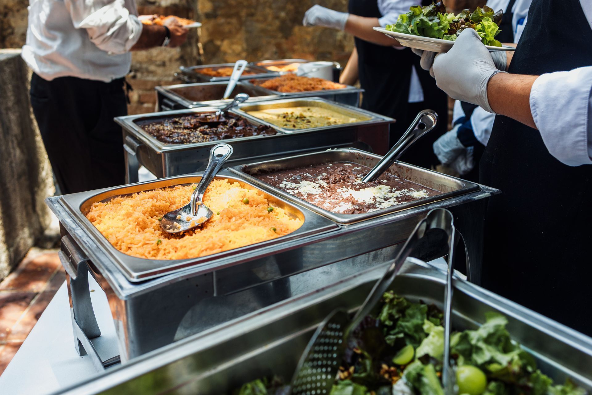 Catering buffet with trays of food being served, including orange casserole and greens, at an outdoor event