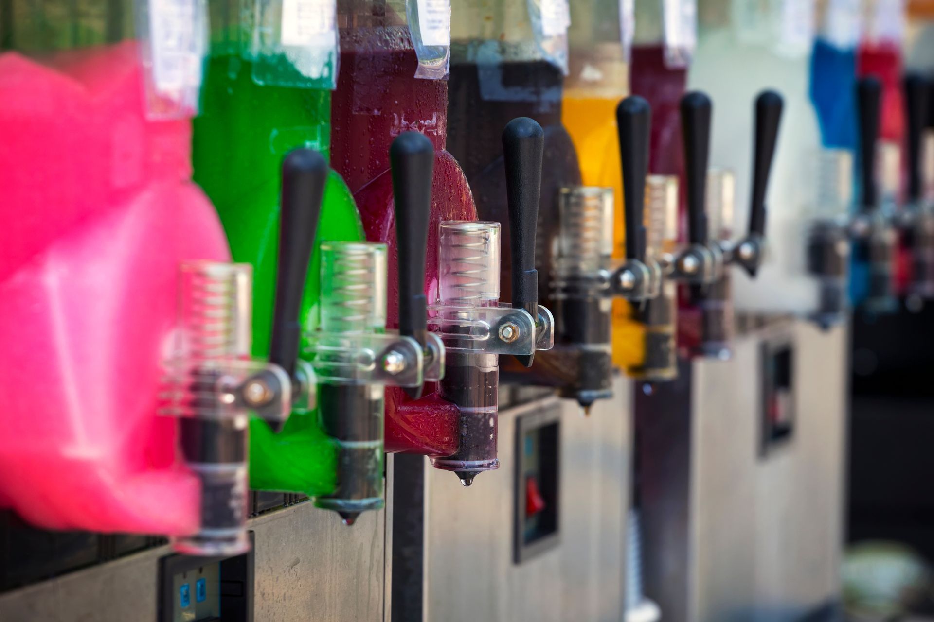 Rows of colorful beverage slush machines with clear handles and metal dispensers in a shop