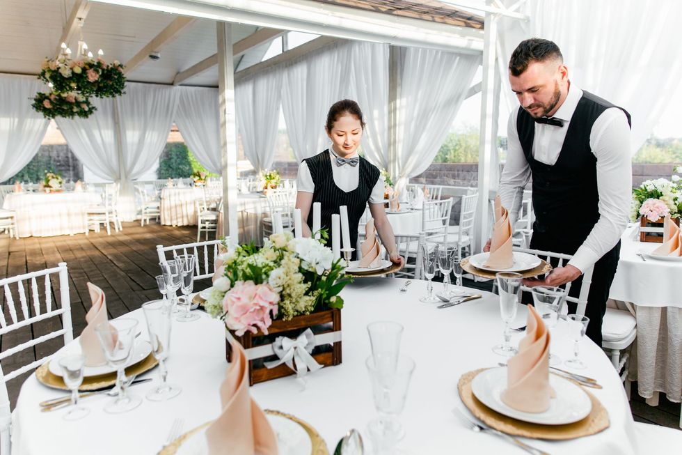 Event staff setting tables in a white tent with floral centerpieces and gold-accented place settings Event staff setting tables in a white tent with floral centerpieces and gold-accented place settings