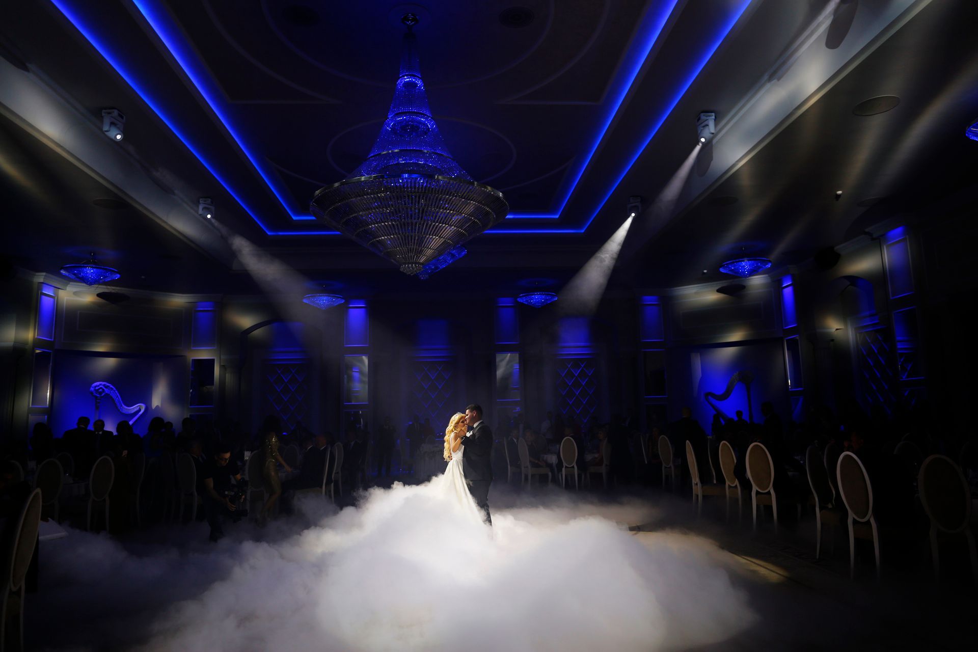 Bride in a white gown standing on a fog-filled stage under blue lights and a chandelier
