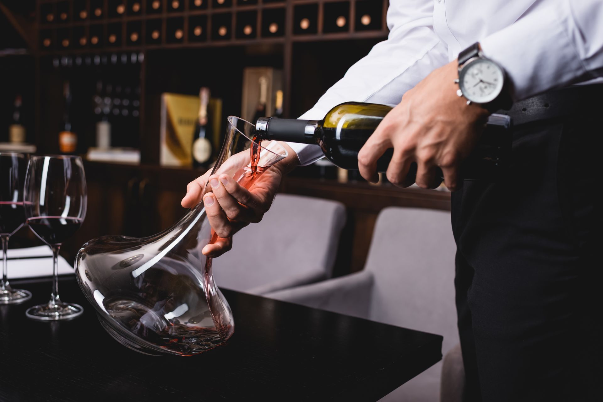 A bartender pours red wine into a glass decanter at a bar table.