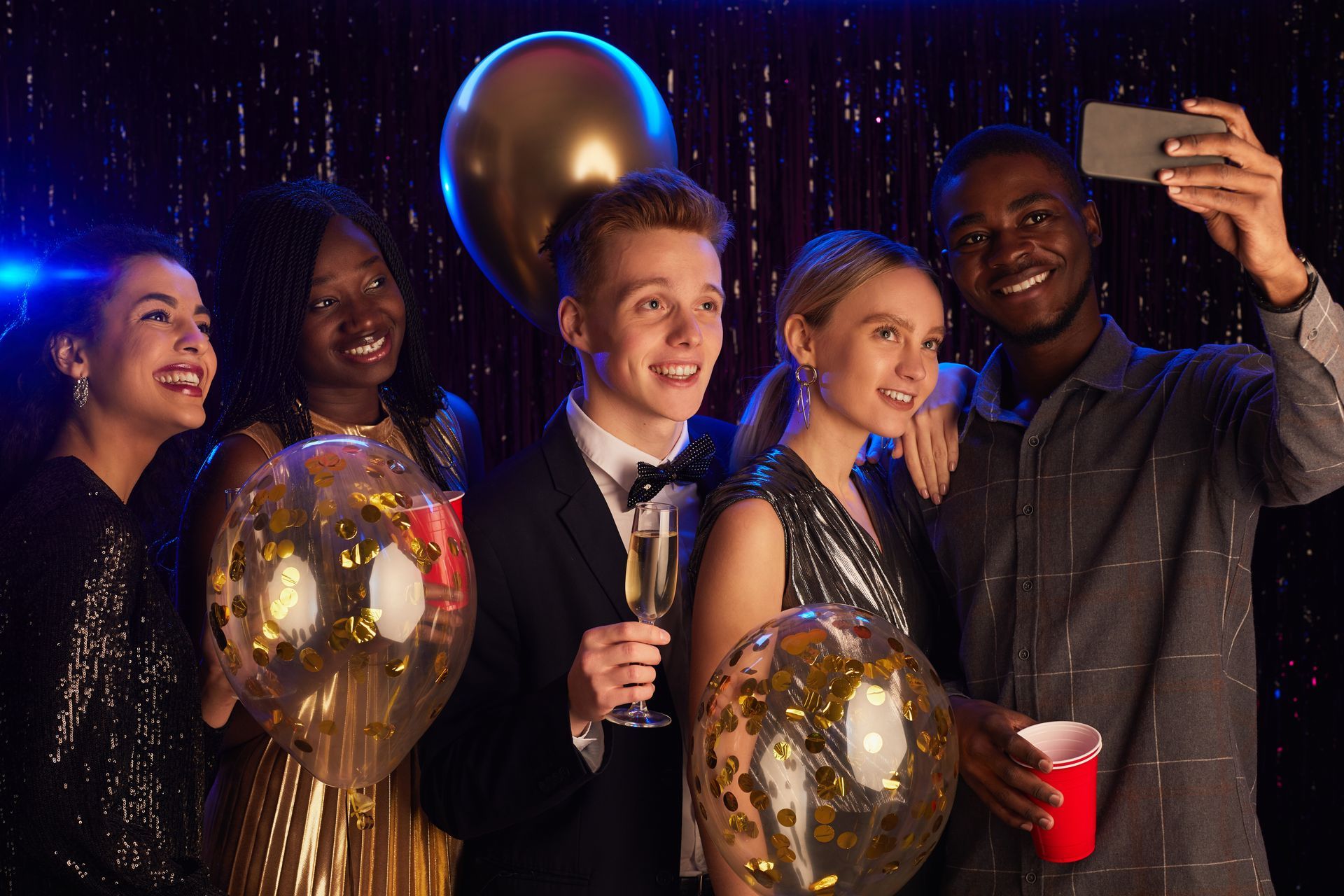 Four friends in party clothes take a selfie with balloons, champagne, and red cups at a festive celebration.