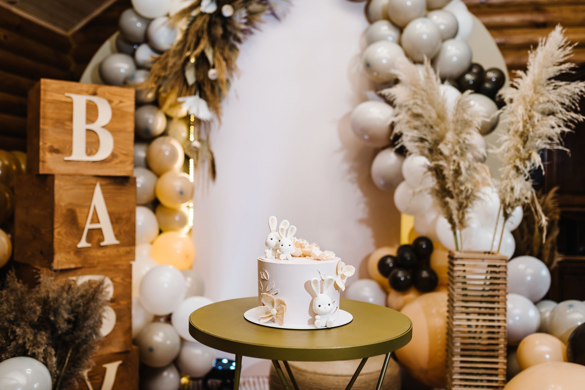 Boho dessert table with a gold cake stand, balloons, pampas grass, and wooden “BA” blocks