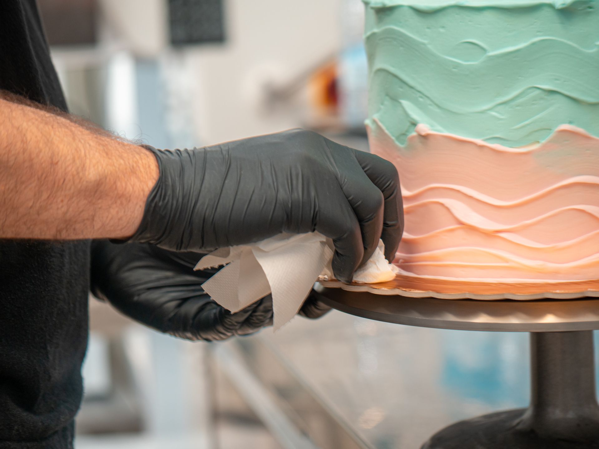 Gloved hands piping white icing onto a pastel pink-and-teal frosted cake on a stand