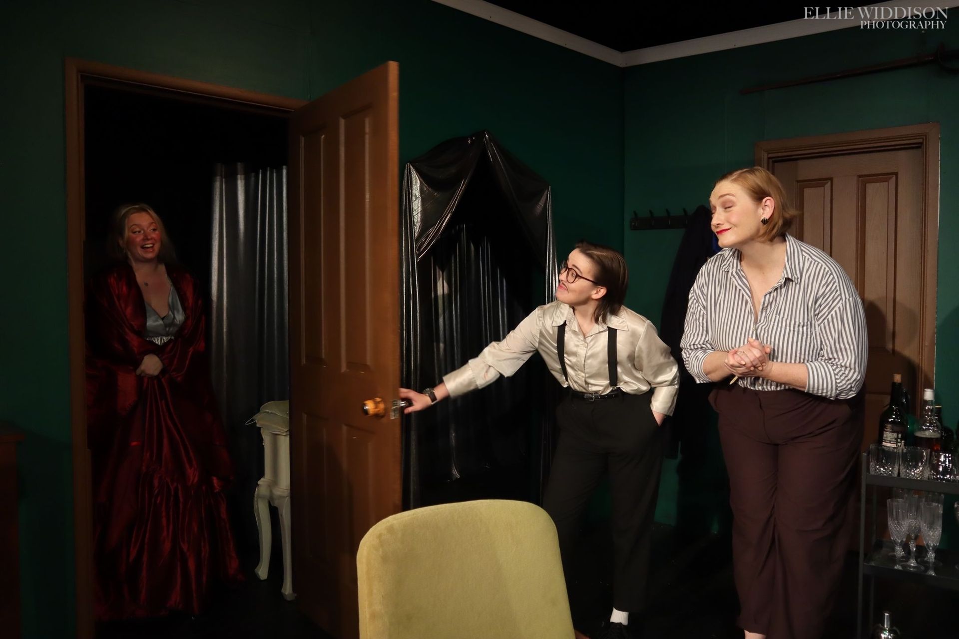 Three people in a green room, one opening a door while two look on, with a figure standing in the doorway.