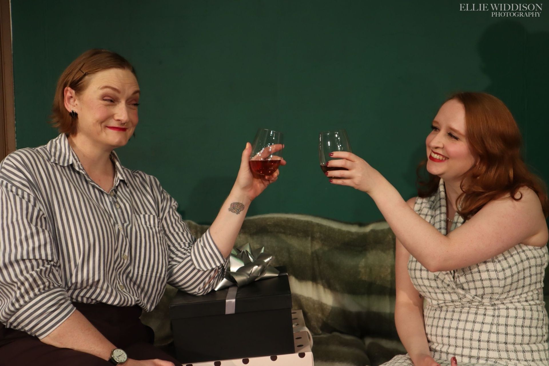 Two people toast drinks while sitting on a couch against a green wall.