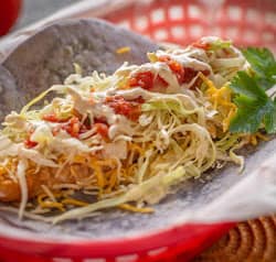 Taco in a red basket, filled with shredded cabbage, cheese, salsa, and sour cream.