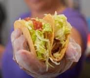 Person holding two tacos with lettuce and tomato in gloved hand.