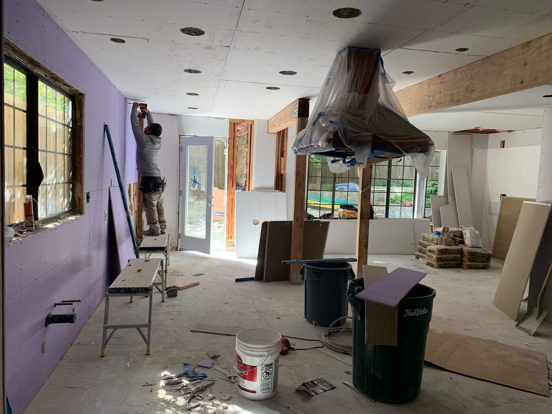 Renovation-in-progress room with drywall, tools, plastic-covered fixture, and a worker on a ladder.