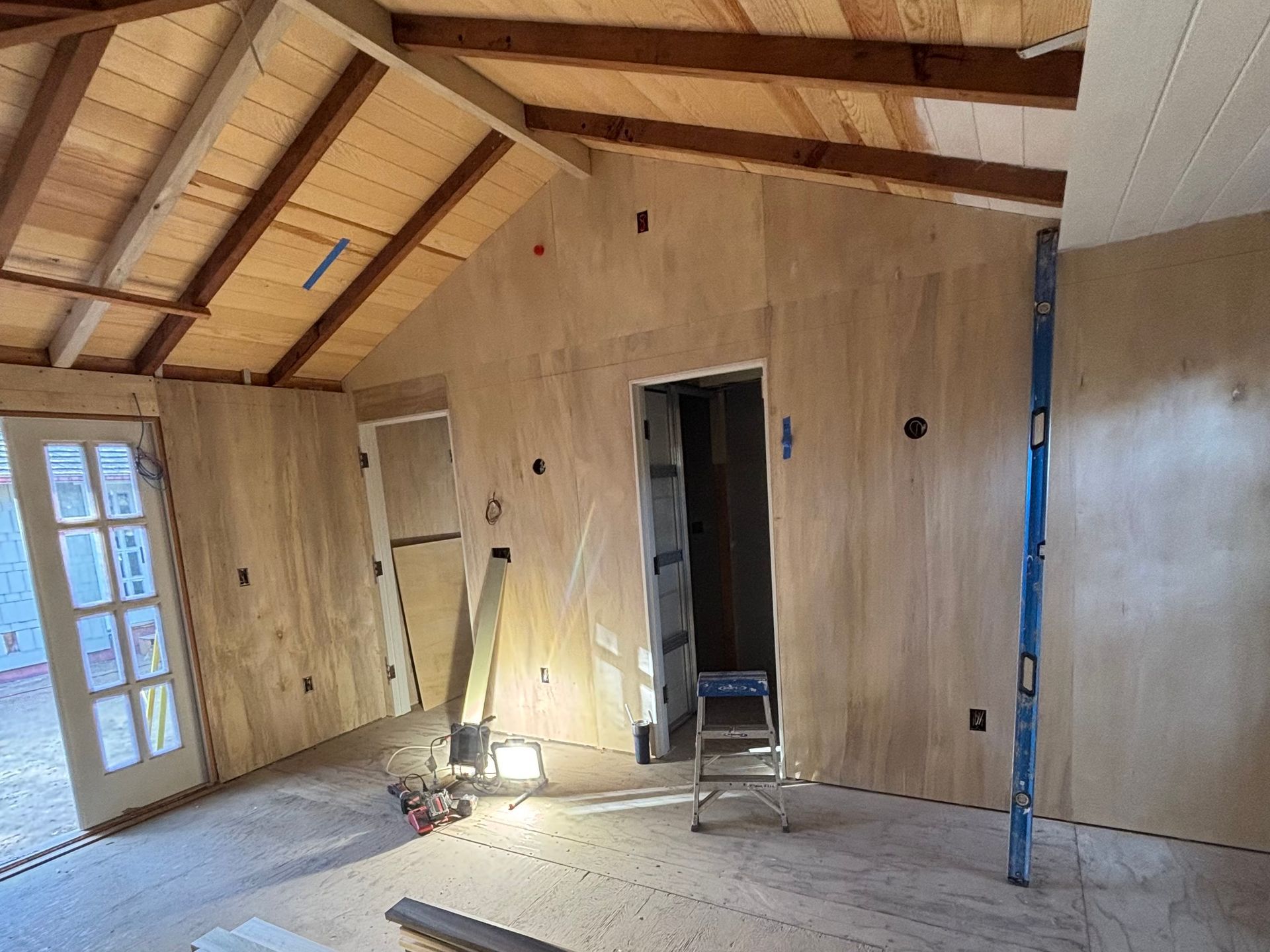 Unfinished room with exposed wooden beams, plywood walls, ladders, and a work light on the floor.