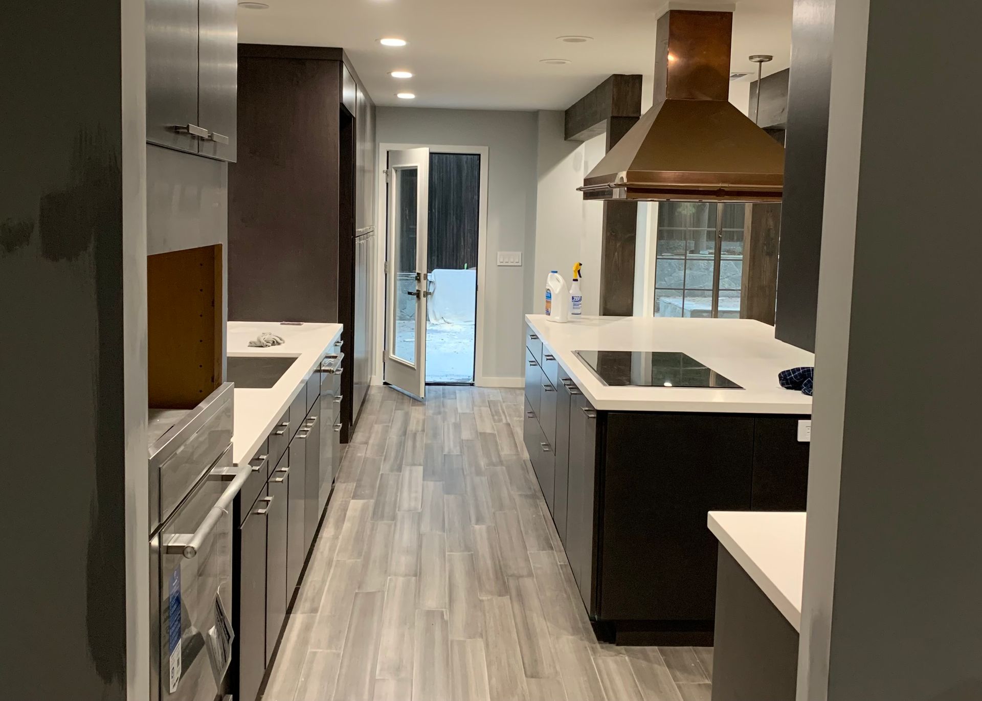 Modern galley kitchen with white cabinets, dark counters, copper hood, and a bright doorway at the end.